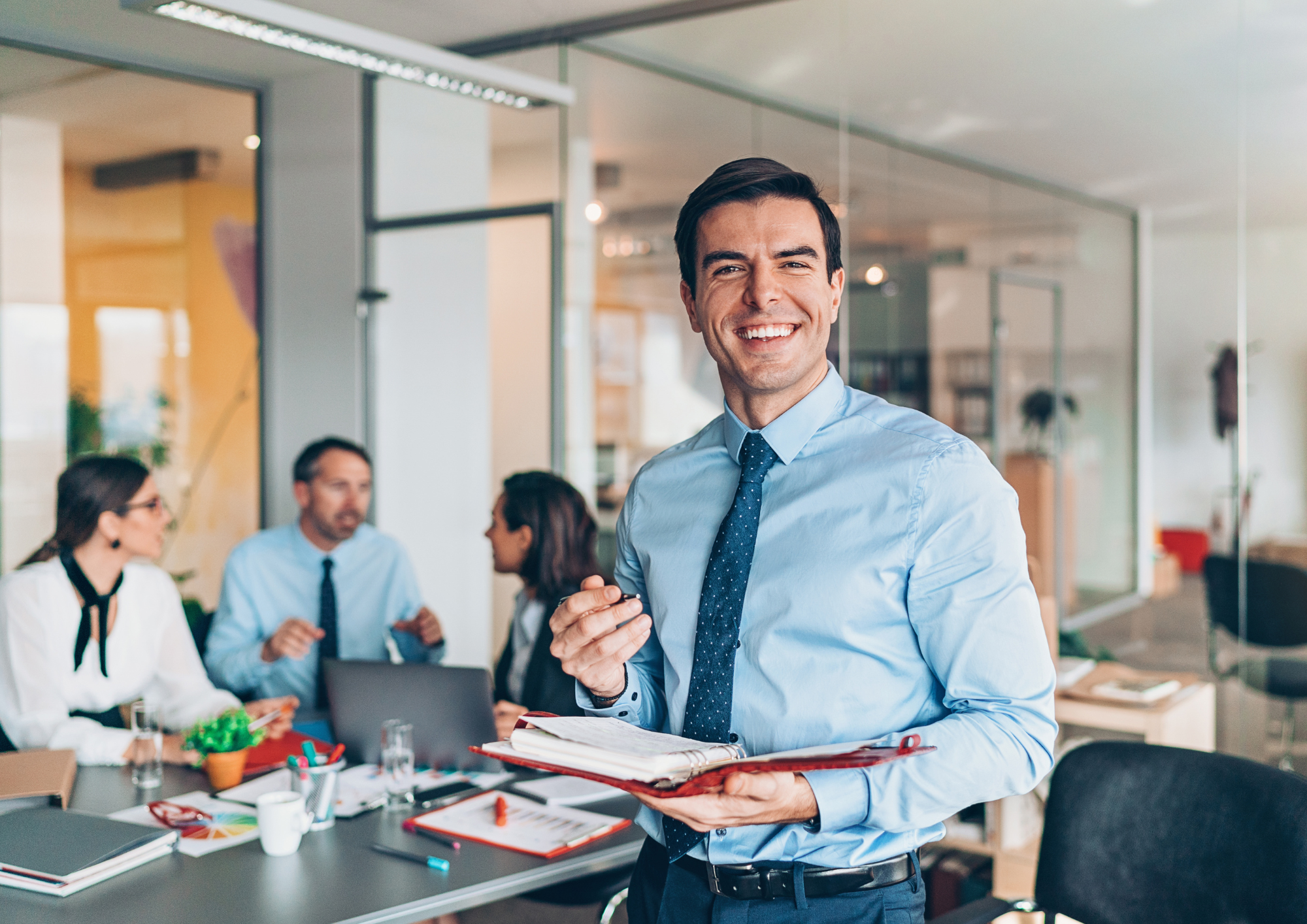 Smiling manager holding a notebook in an office, with colleagues meeting at a table behind him